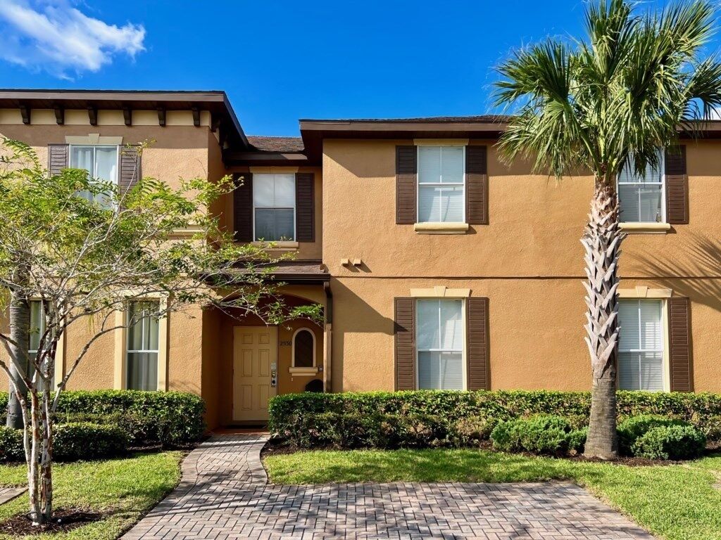 A two-story residential building with a brick pathway leading to the entrance, surrounded by well-maintained hedges and a palm tree.