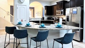 A modern kitchen with a central island, dark cabinets, stainless steel appliances, and blue bar stools. - Luxury 5d Spa Gm Pool Hm Solterra Resort-5165ac 5 Bedroom Home by RedAwning (Davenport)