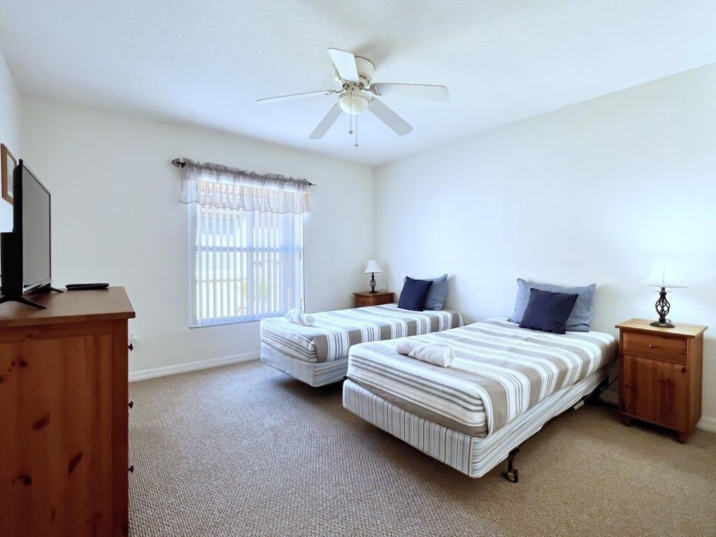 A bedroom with two beds, a ceiling fan, a window with curtains, a wooden dresser, and bedside lamps.