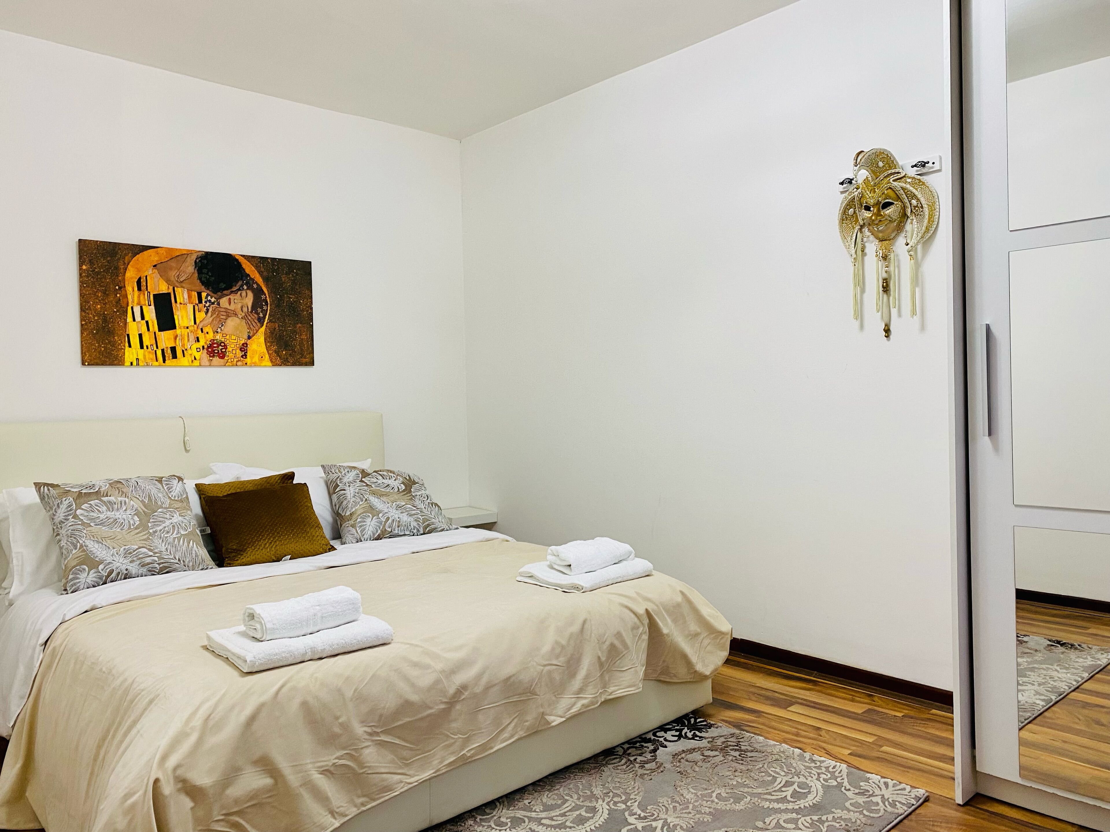 Un lit bien fait, avec du linge de lit blanc, un tableau représentant un couple accroché au mur et un masque décoratif sur la porte.