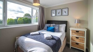 Een net opgemaakt bed met een deken in blauw en wit, een bedlampje en een raam met gordijnen.