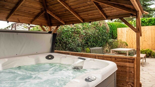 Een hottub onder een houten pergola, met een houten omheining en groen in de achtergrond.