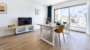 Ett modernt vardagsrum med en platt-TV, trägolv, ett matbord med stolar och ett stort fönster med utsikt över staden.