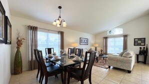 A dining area with a table set for four, surrounded by chairs, a chandelier, and a sofa in the background.