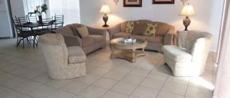 A living room with a sofa, armchairs, a coffee table, and a dining table with chairs. There are two wall-mounted lamps and framed pictures on the wall.