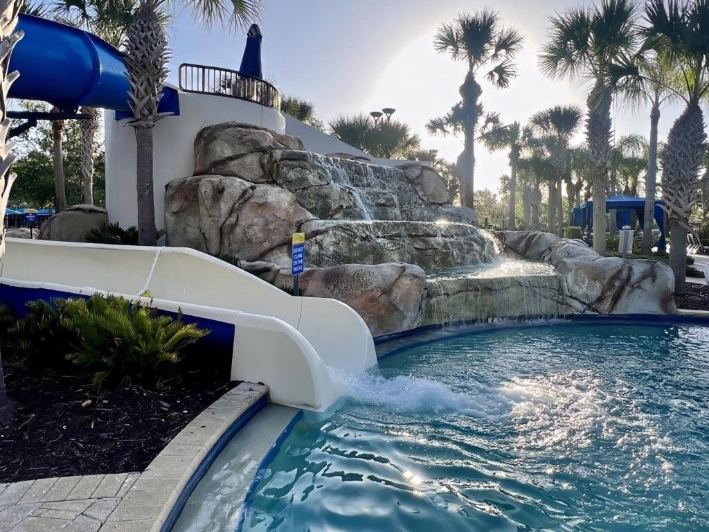 A pool with a water slide, palm trees, and a rocky waterfall feature.