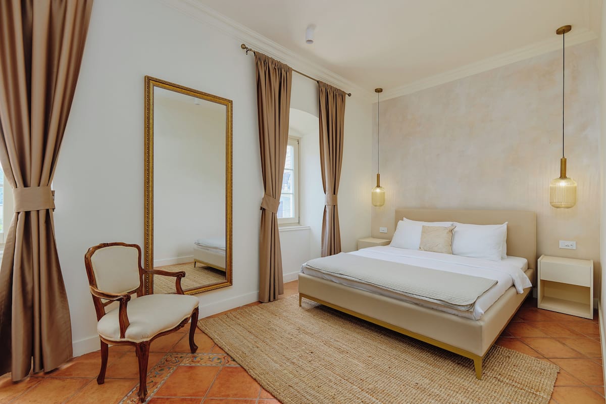 Ein Schlafzimmer mit einem großen Bett, einem Sessel, einem Spiegel und Vorhängen.