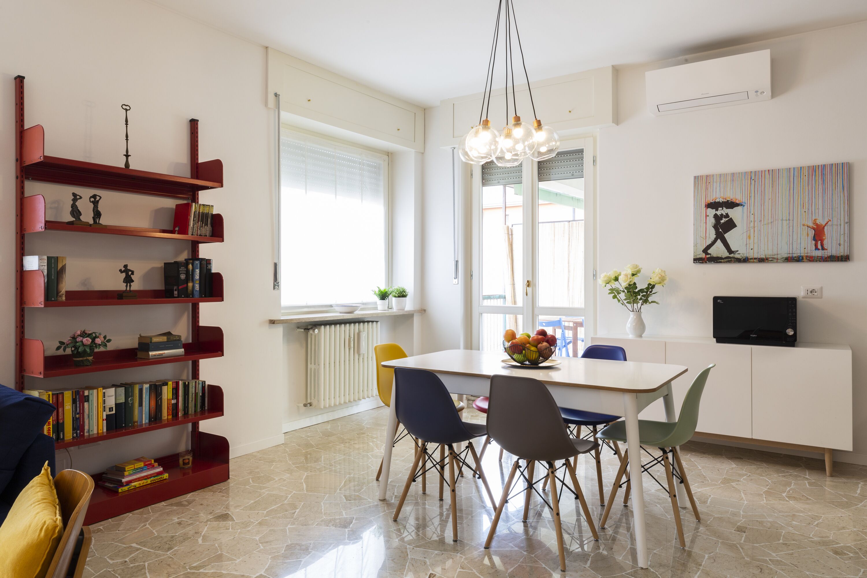 A modern dining area with a white table, colorful chairs, a red bookshelf, and a painting on the wall.