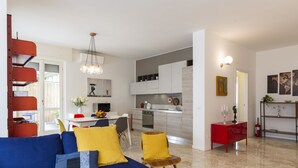 A modern living room with a blue sofa, yellow cushions, and a red side table. A kitchen with white cabinets and a dining area with a table and chairs is visible. - notaMI - CASA RELAX - Central Station (Milan)