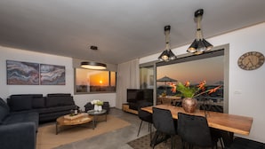 Un salon moderne avec un canapé, une table basse et un coin repas. Une grande fenêtre offre une vue sur le coucher du soleil.