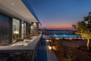 A modern outdoor pool area with stone walls, seating, and a view of the city at dusk. - Villa Karydia with private pool (Kos)