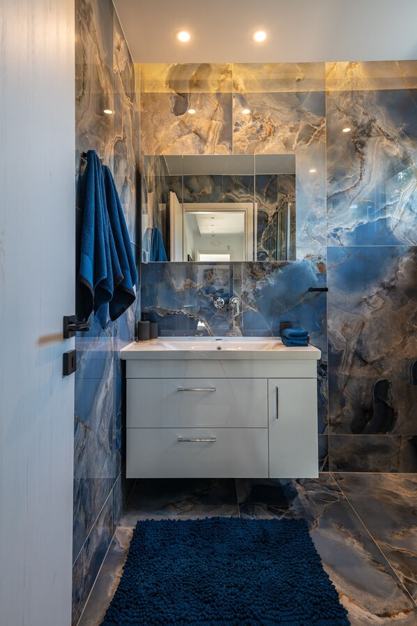 A modern bathroom with marble walls, a white vanity, a blue rug, and a mirror above the sink. - Villa Karydia with private pool (Kos)