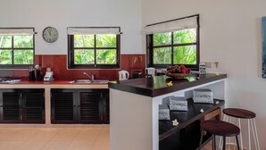 A kitchen with a central island, two windows with curtains, a clock, and a view of greenery outside. - Gorgeous beachfront Villa Jasmine, Lovina! (Kecamatan Banjar)