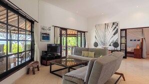 A spacious living room with a sofa, coffee table, and a TV. Large windows offer a view of the outdoors. - Gorgeous beachfront Villa Jasmine, Lovina! (Kecamatan Banjar)