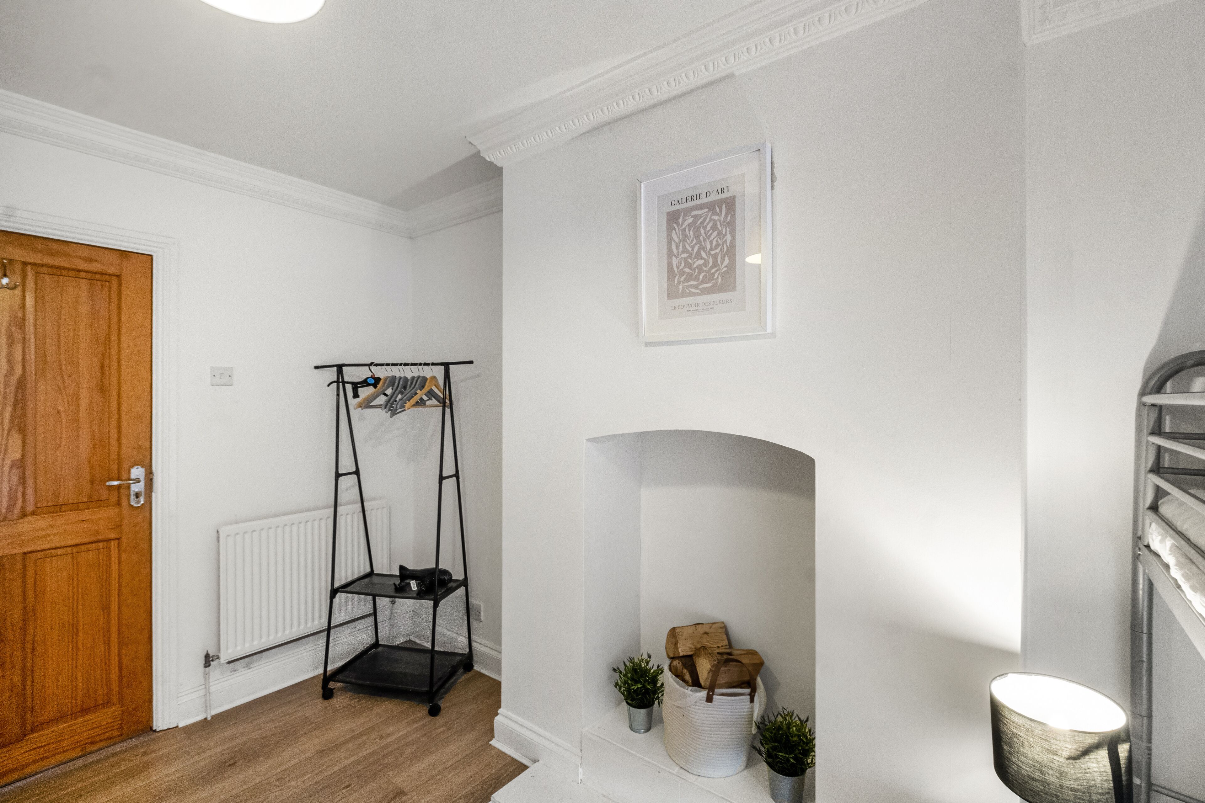 A hallway with a wooden door, a radiator, a black metal shelf, a fireplace with logs, and a framed artwork on the wall.