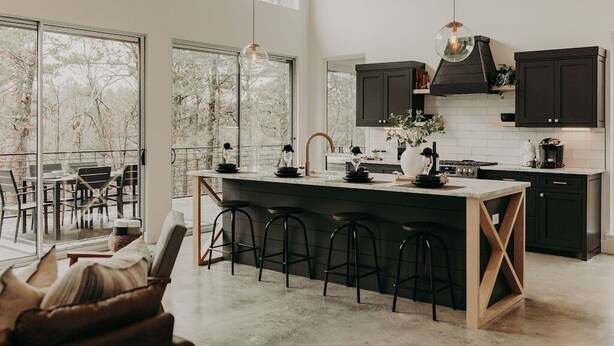 A modern kitchen with a large island, dark cabinetry, and large windows overlooking a snowy landscape. - Secluded 2-King Suite Cabin w/ Hot Tub, Fireplace & Forest Views (Broken Bow)