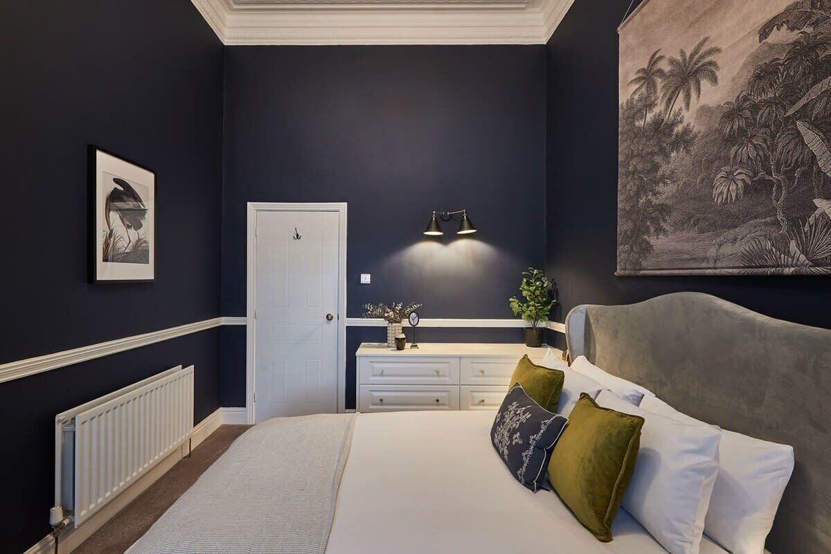 Une chambre à coucher comprenant un lit, une table de chevet, une commode, une porte et un luminaire fixé au mur.