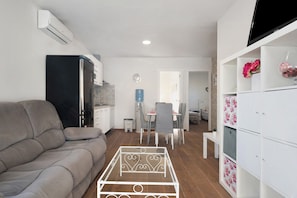 A living room with a sofa, a dining area with chairs and a table, a kitchen with a refrigerator, and a TV mounted on the wall.