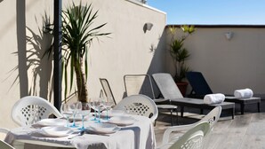 Un espace de restauration en plein air avec une table dressée pour quatre personnes, entourée de chaises blanches, et un coin salon avec des chaises longues et un pot de fleurs.