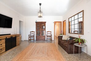 A living room with a TV, wooden cabinet, two chairs, a sofa, a rug, a small table with a plant, and a mirror on the wall. - Exclusive Villa for Groups in Chiclana de la Frontera (Chiclana de la Frontera)