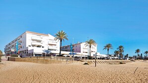 A beachfront hotel with multiple floors, palm trees, and a sandy beach in the foreground.