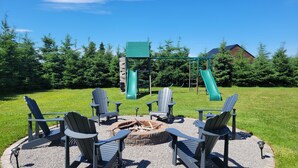 En rund sittplats med Adirondack-stolar runt en eldstad, en lekplats med rutschkanor i bakgrunden och ett hus som syns bortom träden.