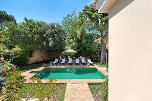 Una piscina con sillones de descanso, rodeada de vegetación y una pérgola.