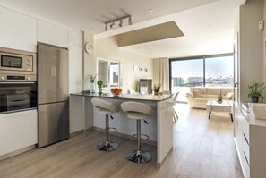Une cuisine moderne dotée d’un four et d’un réfrigérateur encastrés, d’un îlot central avec des tabourets de bar, d’un coin repas avec un canapé et une table basse, et d’une grande fenêtre offrant une vue sur la ville.