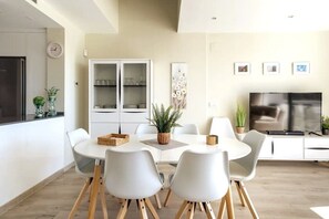 Une cuisine moderne avec des meubles blancs, une table à manger avec quatre chaises, un téléviseur et une horloge au mur.