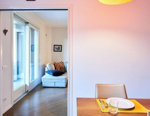 Ein modernes Wohnzimmer mit einer Couch, einem Esstisch für zwei Personen und einem großen Fenster mit Tageslicht.
