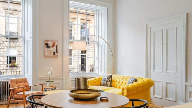 A room with a yellow sofa, a round wooden table, a radiator, a window with a view of buildings, and a door with paneling. - Drake Suite - City Centre 1 Bed Apartment (Edinburgh)