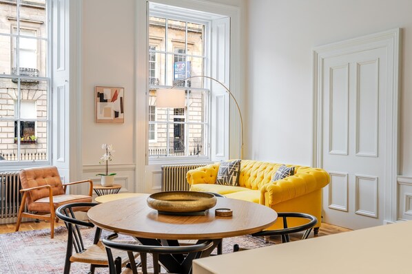 A room with a yellow sofa, a round wooden table, a radiator, a window with a view of buildings, and a door with paneling.