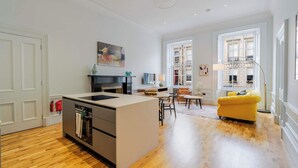 A modern kitchen with a central island, a dining area with a yellow sofa, and a view of the city through large windows. - Drake Suite - City Centre 1 Bed Apartment (Edinburgh)