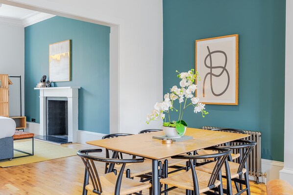 Un coin repas avec une table et des chaises en bois, une cheminée blanche et un mur de couleur turquoise orné d’une œuvre d’art abstraite encadrée.
