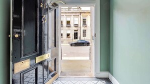 A black door with a brass handle and a number plate, leading to a street with a parked car and a building with balconies.