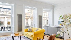 A bright living room with a yellow sofa, a dining table, and a view of the city through large windows.
