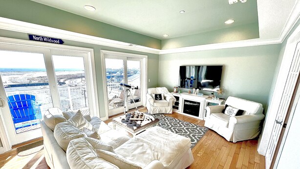 A spacious living room with a large sofa, a coffee table, and a flat-screen TV. The room has large windows offering a view of the city.