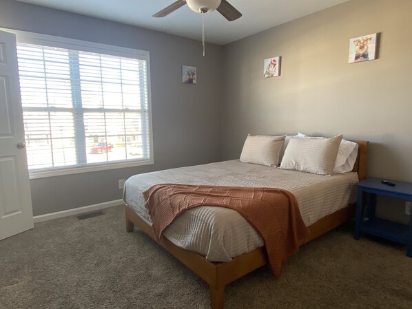 A bedroom with a bed, a ceiling fan, a window with blinds, and a small bedside table.