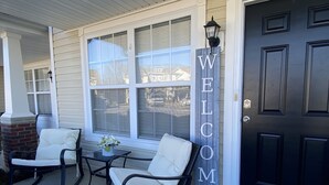 Eine Veranda mit zwei Schaukelstühlen und einem kleinen Tisch, eine schwarze Tür mit einem 'WELCOME'-Schild und einer Fußmatte im Bauernhausstil am Eingang.