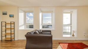 Ein Wohnzimmer mit braunem Ledersofa, Holzboden und drei Fenstern mit Blick auf ein Gewässer.