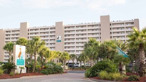 A multi-story hotel with a central entrance, surrounded by palm trees and a parking area.