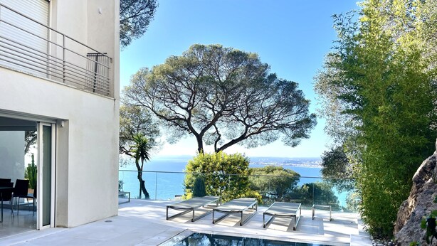 Un espace piscine extérieur moderne, offrant une vue dégagée sur la mer et entouré d’arbres et d’un bâtiment.