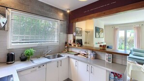 A modern kitchen with white cabinets, a built-in oven, and a large window with blinds. - Villa des Doudoux (Andernos-les-Bains)