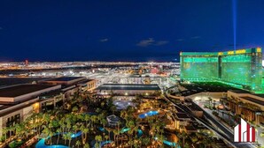 Vista nocturna de un complejo hotelero con varias construcciones, incluyendo un MGM Grand destacado con señalización iluminada. Hay varias piscinas rodeadas de palmeras y un estacionamiento bien iluminado.
