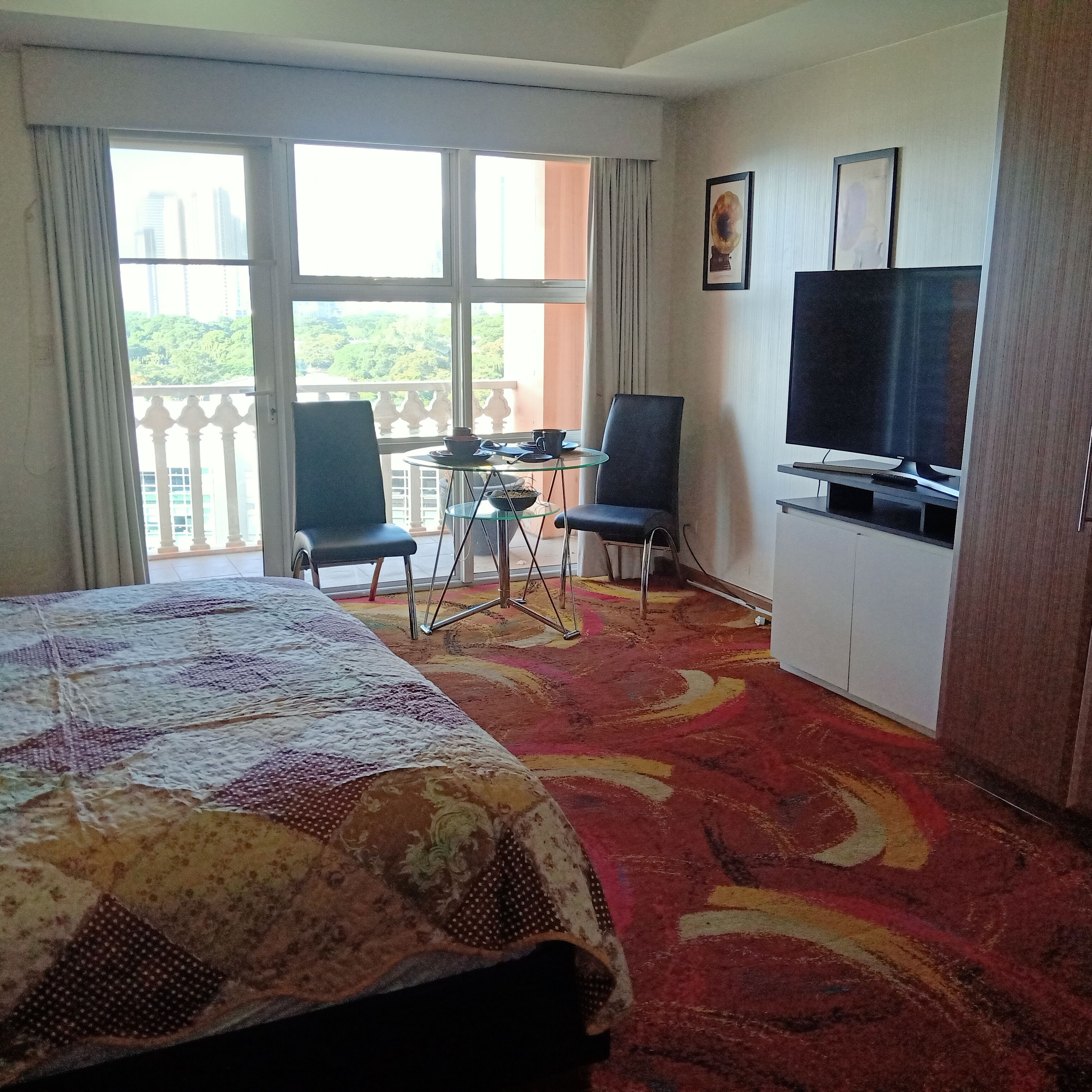 Une chambre d’hôtel comprenant un lit, deux chaises, une petite table, un téléviseur et un balcon avec vue sur la verdure.