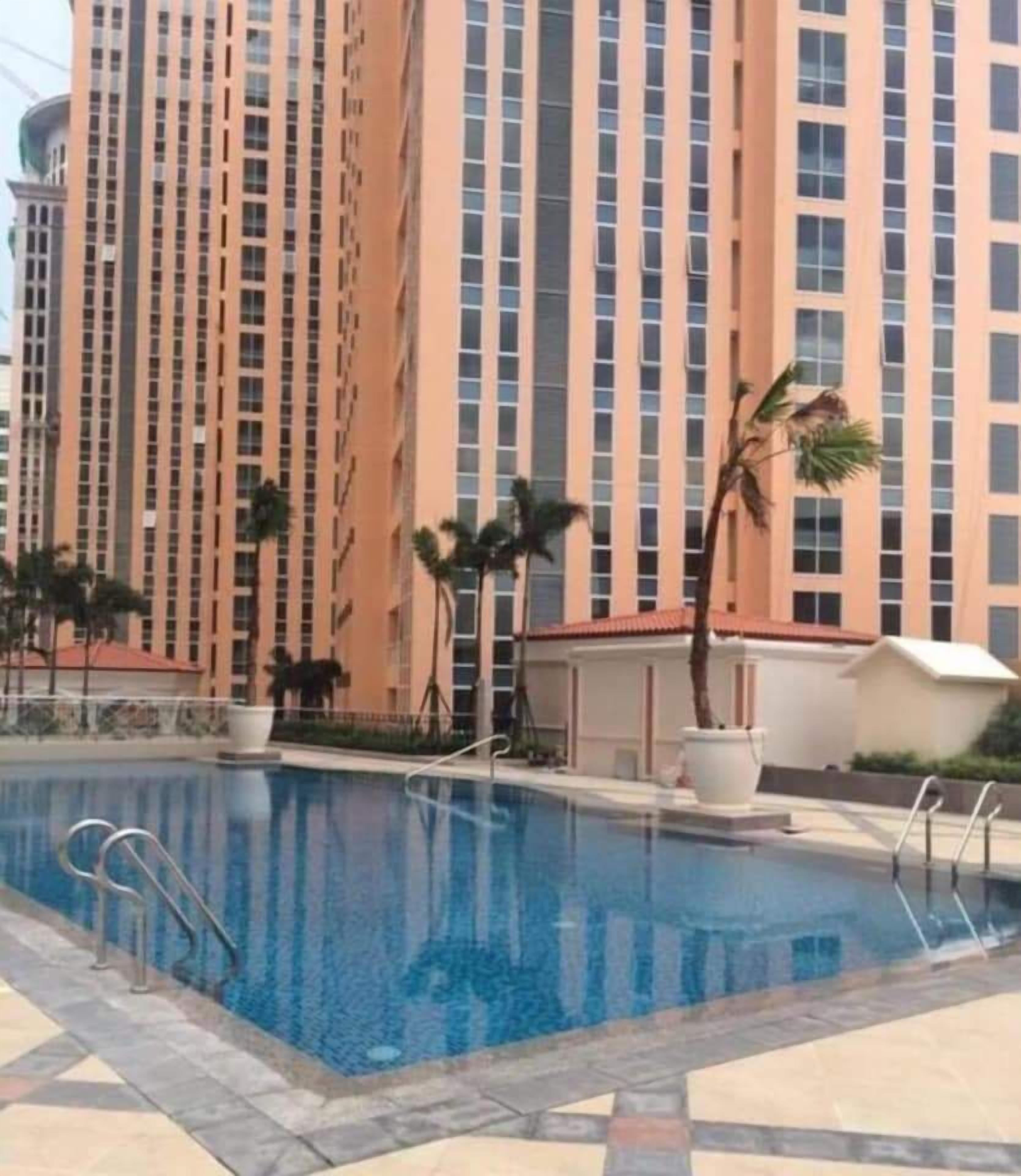 Una piscina con superficie de agua azul cristalina, rodeada por una terraza con baldosas. Al fondo, un edificio residencial alto con fachada color beige y numerosas ventanas.