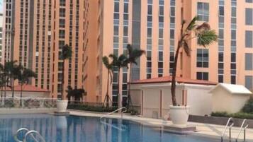 Una piscina con superficie de agua azul cristalina, rodeada por una terraza con baldosas. Al fondo, un edificio residencial alto con fachada color beige y numerosas ventanas.