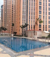 A swimming pool with a clear blue water surface, surrounded by a tiled deck. In the background, there is a tall residential building with a beige facade and numerous windows.
