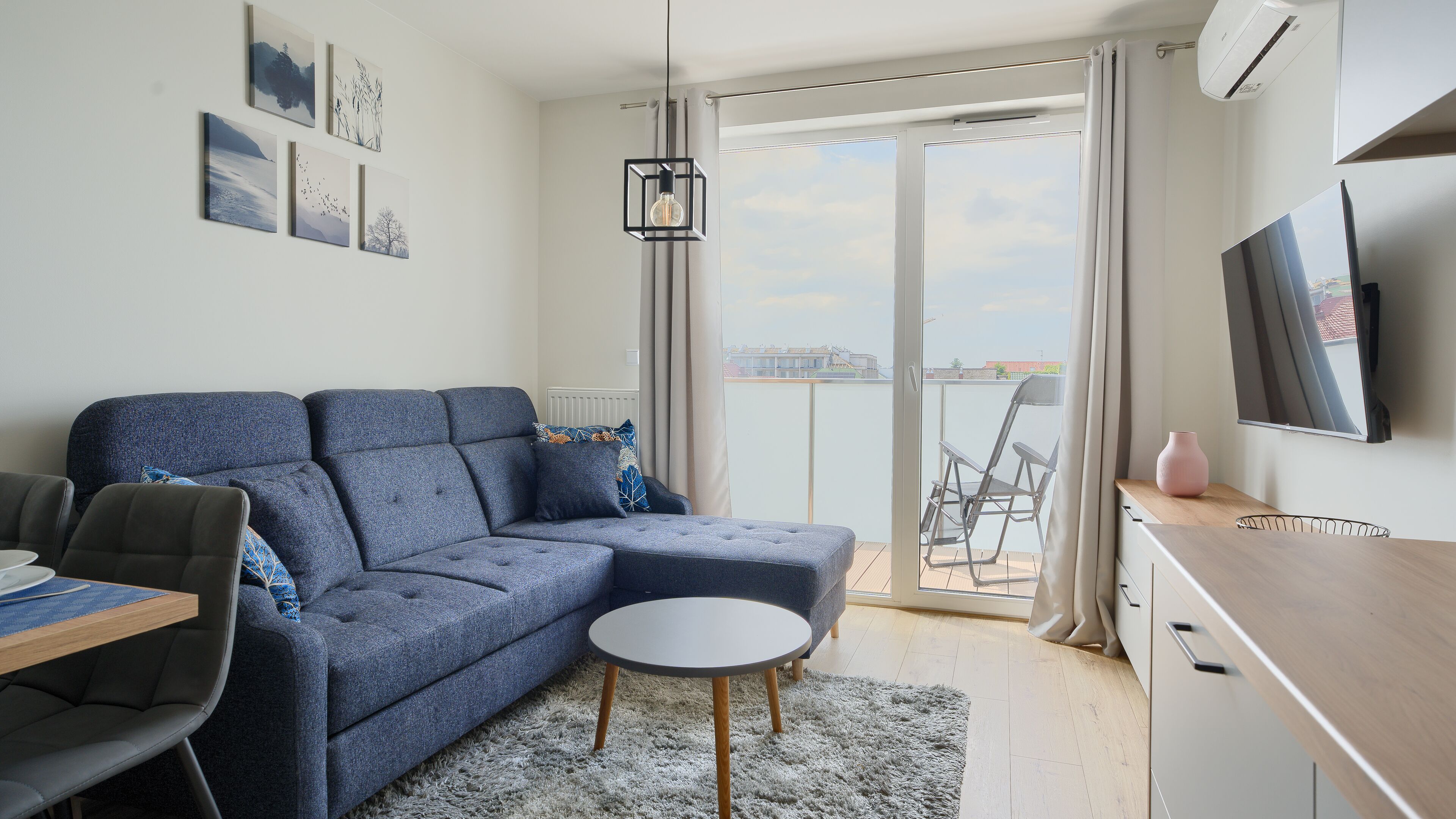 A modern living room with a grey sectional sofa, a round coffee table, a dining area with a wooden table and chairs, a TV mounted on the wall, and a balcony with a view of buildings.