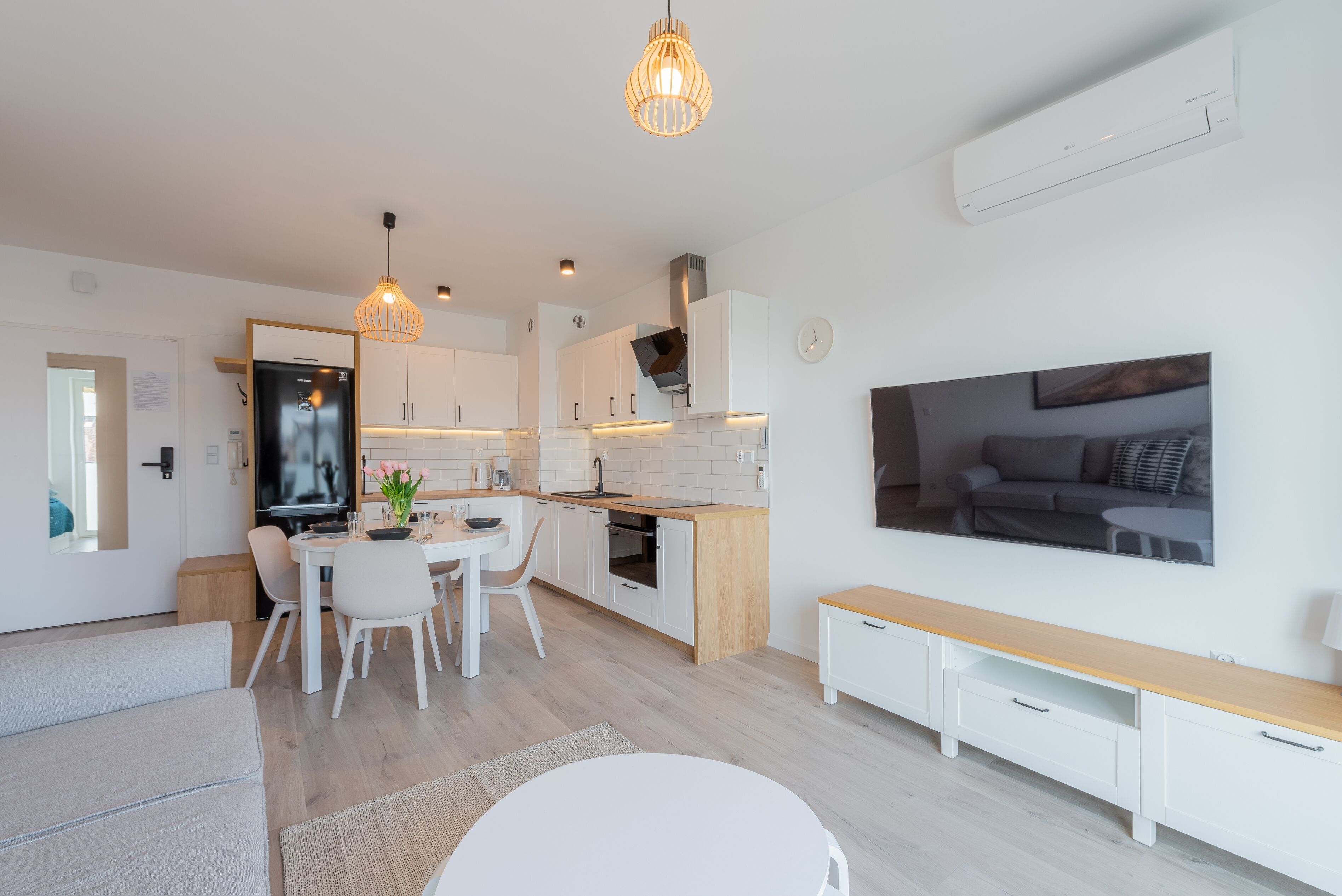 A modern kitchen with white cabinets, a dining area with a white table and chairs, and a living area with a sofa and a TV.